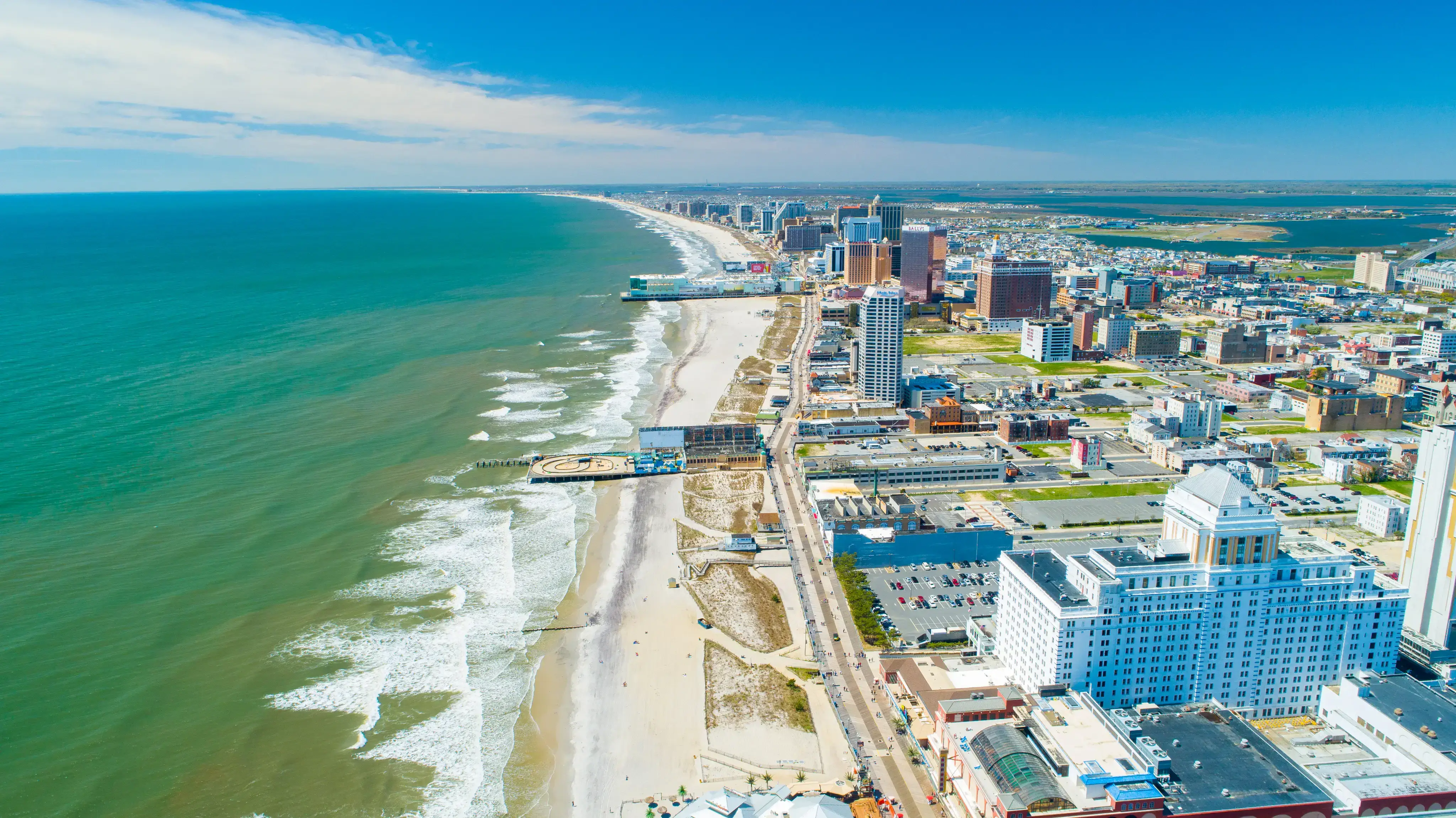 Aerial view of a coastal city with a sandy beach, ocean waves, piers, and a skyline of tall buildings under clear blue skies.