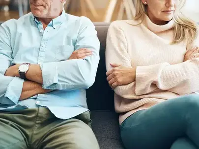 A man and woman sit side by side on a couch with arms crossed, facing away from each other, appearing upset or distant.