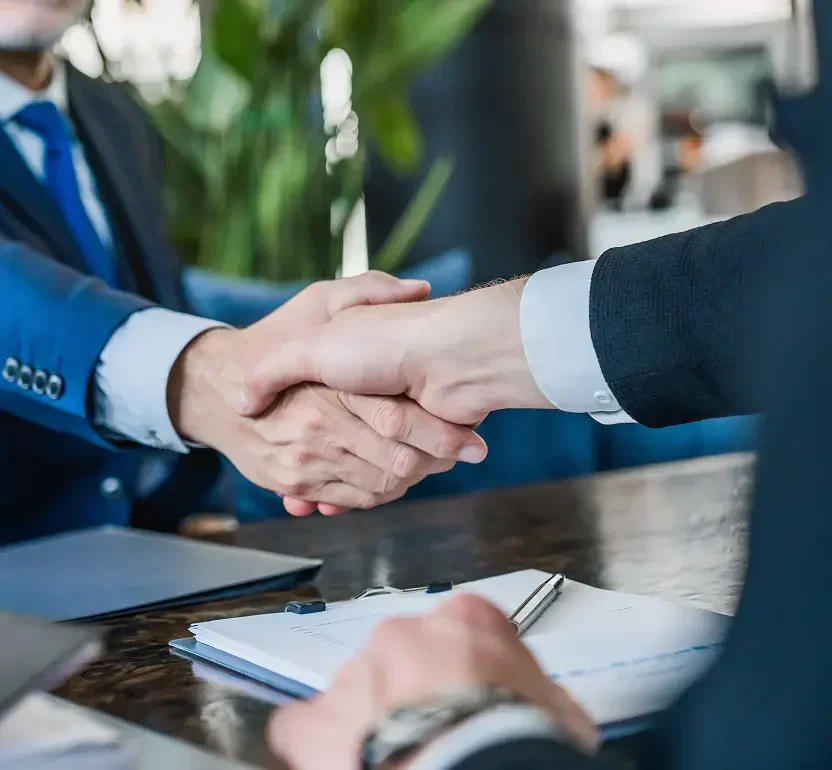 Two business professionals shaking hands over a desk with documents and a pen, symbolizing agreement or partnership.