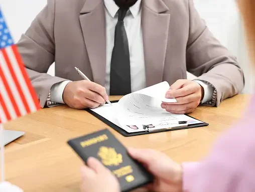 Person in a suit reviewing documents on a clipboard while another holds a U.S. passport at a desk with an American flag in the background.